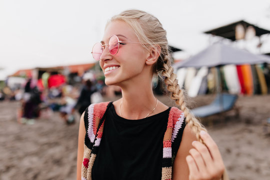 Winsome Tanned Girl Posing With Smile At Beach. Outdoor Portrait Of Blonde Woman In Pink Glasses Playing With Her Braids.