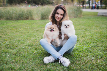 A young pretty woman sits on the green grass and holds her beloved Pomeranian dogs. Spring day. Two Pomeranian dogs with their owner 
on a walk. Selective focus.
