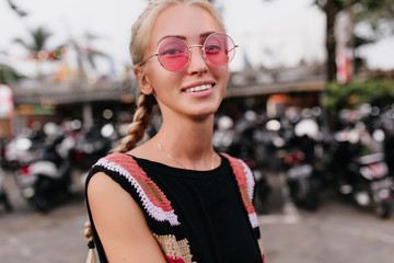 Close-up shot of inspired girl with blonde braids. Photo of excited tanned female model in pink sunglasses and knitted attire posing on blur background.