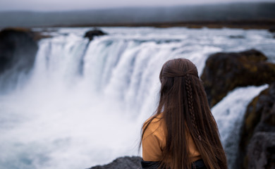 Young woman sitting at the Godafoss waterfall in Iceland. Godafoss means the waterfall of the gods in icelandic.