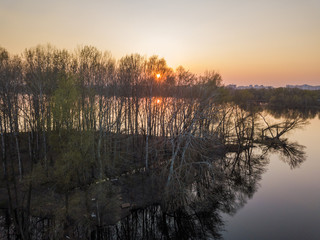 The rays of the setting sun through rare trees on the island in the lake. Aerial drone view.