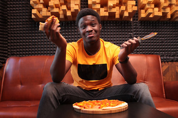 A cheerful black guy is sitting on the couch with a slice of pizza and a pizza knife. Photo in the interior. The concept of a good mood. Copy of the space.