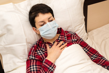 Sick woman in medical mask lying in bed feeling chest pain and panting. Short-haired brunette with symptoms of covid-19 or pneumonia staying at home during coronavirus pandemic.