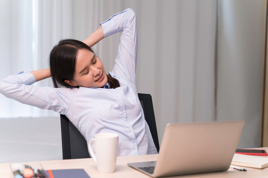 Attractive Asian woman works from home with laptop on desk in bedroom. Young girl raises arms, stretching to relieve pain from office syndrome. Shelter in place from COVID-19 and Stay Home concept.