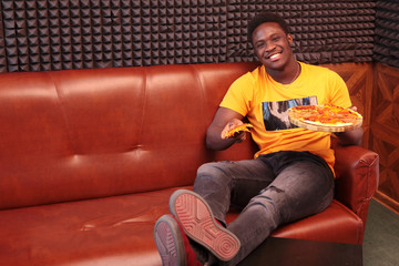 A young African-American man is sitting on a leather sofa with a slice of pizza in his hand. He smiles and looks at the camera. Copy of the space.