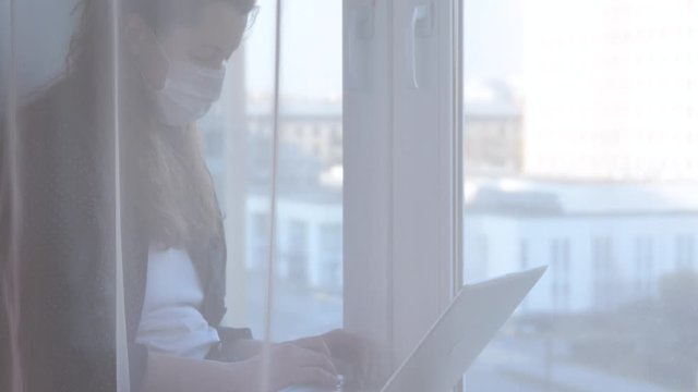 Woman wearing mask and working on laptop from home due to coronavirus self isolation. Sitting on window sill. Authentic home workplace. Handheld shot with gimbal. Coronavirus outbreak 2020.