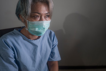 covid-19 virus spreading disease - scared and sad mature woman with grey hair in hospital patient gown and mask infected by coronavirus feeling sick and in panic