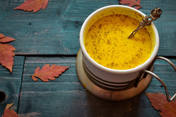 Golden milk in a white cup. Wooden color blue background. Autumn leaves. A healthy drink made from milk, turmeric, cinnamon and honey.
