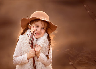 A smiling girl with pigtails in a fur vest and a cute beige hat in early spring with a bouquet of willow branches in her hands 