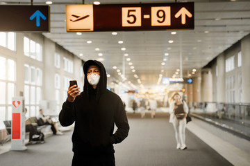 Men in respirator mask is waiting next plane at the airport terminal and using mobile phone. Coronavirus COVID-19 concept.