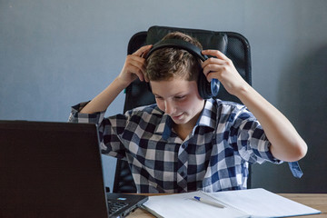  A teenager in a checked shirt and studying at home on a laptop. He sits at the table and puts on his headphones