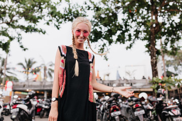 Fototapeta na wymiar Shapely girl with long braids posing with smile on street background. Outdoor portrait of wonderful blonde female model in black dress and pink sunglasses.