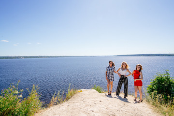 Shoot of three beautiful girls outdoors by the river. Female friends relaxing by the river and smiling. Girlfriends are stand on the pier, joy, fun, hands up. Girls with drinks in dresses. Summer