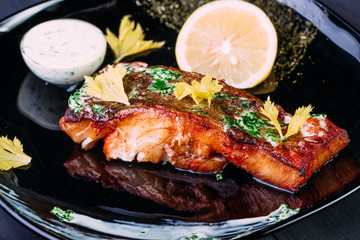 One piece of baked salmon grilled pepper, lemo and white souse on a black plate. wood blue background