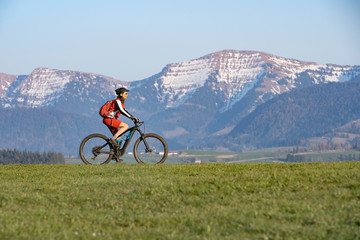 pretty senior woman riding her electric mountain bike in early springtime in the Allgau mountains near Oberstaufen, in warm evening light below the spectacular snow capped mountains of Nagelfluh chain