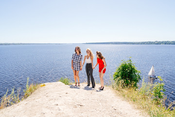 Shoot of three beautiful girls outdoors by the river. Female friends relaxing by the river and smiling. Girlfriends are stand on the pier, joy, fun, hands up. Girls with drinks in dresses. Summer
