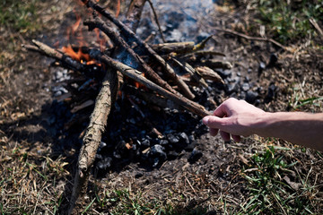 Fototapeta premium Ternopil/Ukraine-Apr 04 2020: Close-up picture of bonfire, made of dry branches, outside on green grass at sunny weather in summer. Hand, holding wooden stick. fixing fireplace in forest.