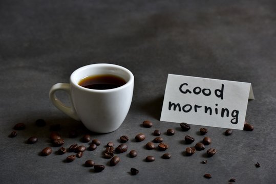 A fresh Cup of brewed coffee on a dark background and a greeting note in the notebook Good Morning 