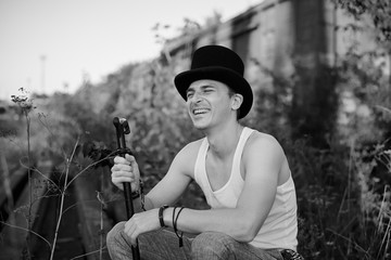Young handsome mad, wearing grey pants and white t-shirt, holding black classic hat, sitting on abandoned railway track by old train, smiling, laughing. Black and white picture of artist, posing. - Powered by Adobe