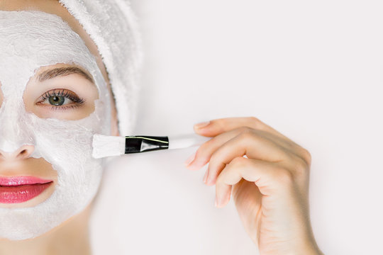Cropped image of beautiful and attractive young woman with hair wrapped in white bath towel, applying mud mask with brush, lying on isolated white background. Professional face skin beauty treatment