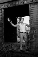 Full-length portrait of young man, wearing grey pants, white top, standing inside old railway carriage, leaning on doors. Black and white picture of creative man on abandoned train area. Art-house.