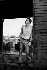 Full-length portrait of young man, wearing grey pants, white top, standing inside old railway carriage, leaning on doors. Black and white picture of creative man on abandoned train area. Art-house.