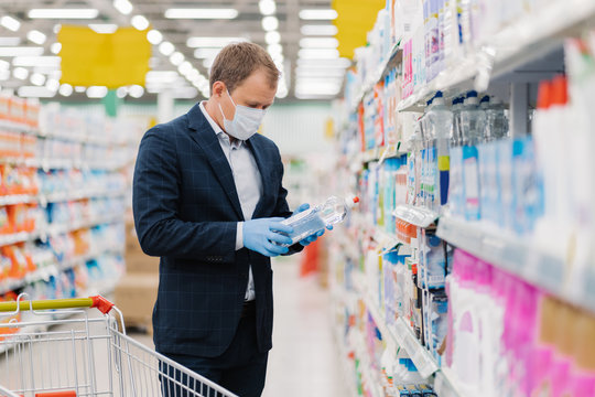 Shot Of Adult Man Chooses Detergent In Housekeeping Store, Reads Label And Instruction How To Use Product, Wears Medical Mask And Gloves During Coronavirus Pandemic, Avoids Risk To Catch Virus