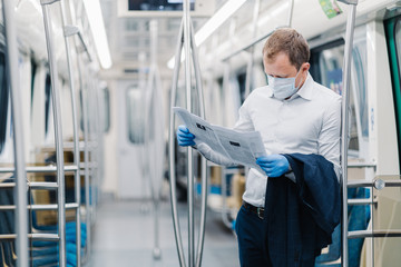 Horizontal shot of adult man wears formal clothes, protective medical mask and gloves, reads press, finds out news during virus outbreak, commutes to work in public transport. Coronavirus, Covid-19