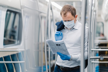 Naklejka na ściany i meble Coronavirus pandemic, Covid-19, quarantine measure. Serious adult man in white shirt, holds black jacket, concentrated in newspaper, wears protective mask and gloves, poses at metro carriage