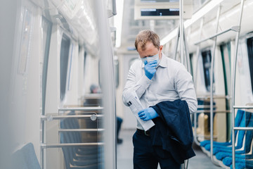 Sick man commuter sneezes constantly, has symptoms of coronavirus, wears surgery medical mask and gloves, holds jacket and newspaper, stands in public transport, has responsible social behavior