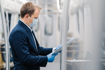 Sideways shot of young elegant man reads newspaper attentively, poses in metro carriage, wears medical mask and disposable rubber gloves from coronavirus. Social distancing, quarantine measures