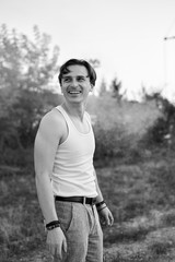 Three-quarter portrait of young man, wearing white top and grey pants,standing in front of bonfire smoke, smiling, laughing. Black and white picture of creative man on abandoned industrial area.