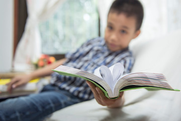 Asian boy holds a book, stretching his hand forward with a cheerful smile. Select focus on the book and blur the background.