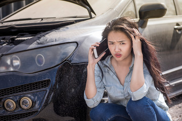 Asian woman sat down near the broken car and opened the hood. She is calling her smart phone and following the mechanic to fix her car. Select focus on her face.