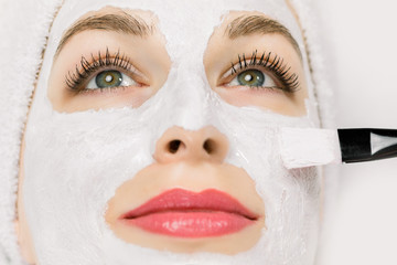 Cropped close up image of pretty young Caucasian woman with towel on head, in health spa center, while white mud clay facial mask is applied on her face. Skin care, beauty concept, facial mask