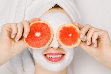 Pretty young girl in bath towel and facial mask with grapefruit slices in front of her eyes. Beautiful woman holding grapefruit slices like eyeglasses, hiding her eyes, lying on white background
