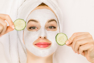 Home spa, skin care concept. Close up top portrait of pretty young woman with towel on her head, white clay mask on her face, holding pieces of fresh cucumber, lying on the isolated white background.