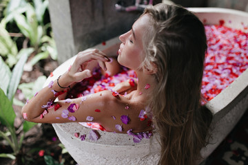 Long-haired tanned woman enjoying spa in weekend. Blonde female model lying in bath with flower petals and chilling with eyes closed.