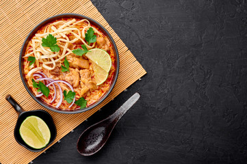 Khao Soi Gai or Thai Coconut Curry Noodle Soup in black bowl at dark slate background. Khao Soy is Thailand cuisine dish with noodles, chicken meat, red chillies paste and spices. Copy space