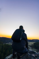 Young man in jacket, pants and backpack sitting on rock looking to valley at sunrise