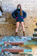 A girl in a protective blue mask and pink gloves, in an old sweatshirt and boots stands on the background of an old brick wall. Stop coronavirus. Chinese concept of pandemic coronavirus. COVID-19