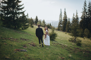 Fototapeta na wymiar Wedding in the mountains. A young couple on a green slope among the pines, a girl in a white dress holds in her hands a bouquet of flowers and greenery with a ribbon