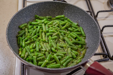 Fresh juicy green french beans are fried in a frying pan on a kitchen gas stove
