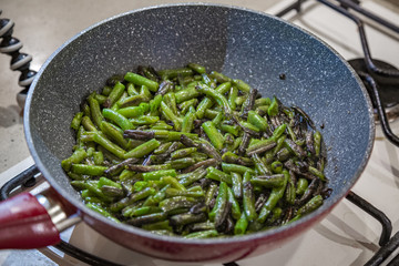 Fresh juicy green french beans are fried in a frying pan on a kitchen gas stove