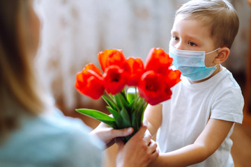 Little son in protective sterile medical mask  congratulates mother and gives a bouquet of flowers tulips. Happy mother's day. The concept of preventing the spread of the epidemic coronavirus.
