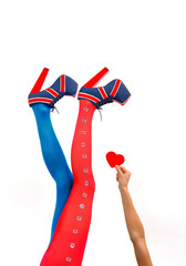 A model's legs are stretched out in a vertical 
form in front of a white background. The woman's 
shoes have a British flag look to them. She 
lifts her arm and holds a red heart in her hand.