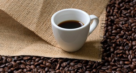 A white Cup of coffee on a background of coffee beans and coarse burlap. Coffee composition.