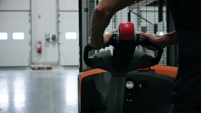 Close up of an operator who is on an electric pallet truck in a warehouse. We can see the hands of the operator who walks. Company and warehouse logistics