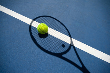 Shadow of a tennis racket and the tennis ball inside it. All on a blue tennis court.