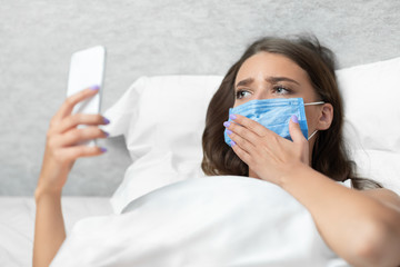 Sad Girl Using Smartphone Wearing Mask Lying In Bed Indoors